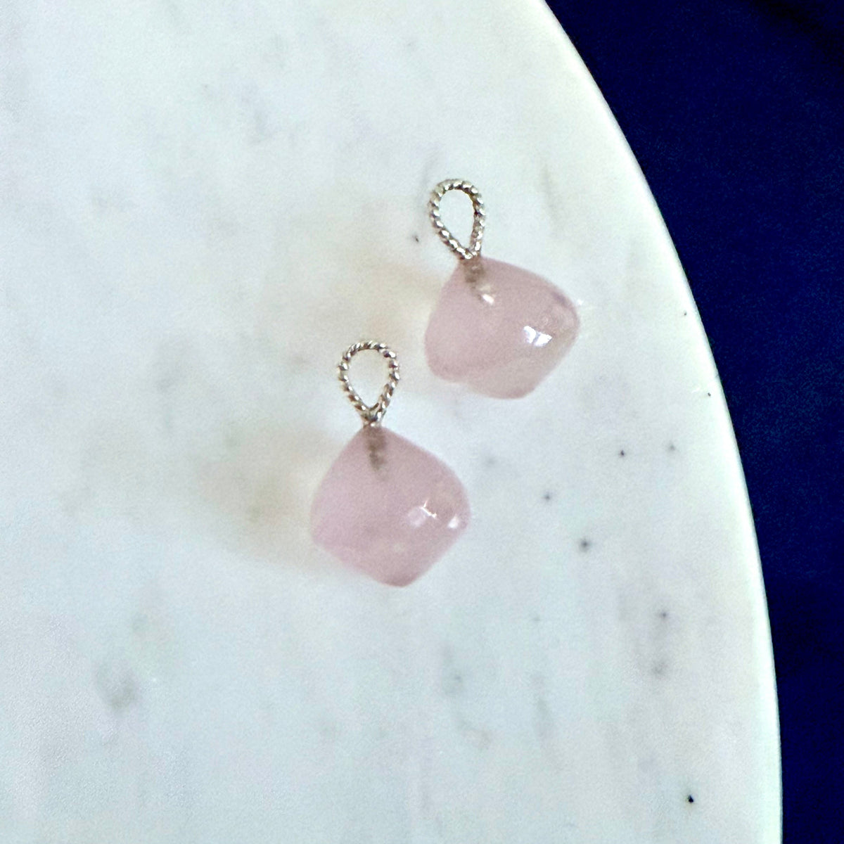 A pair of small drop earrings with light pink stone drops and twisted silver loops, displayed on a white marble surface against a dark blue background.