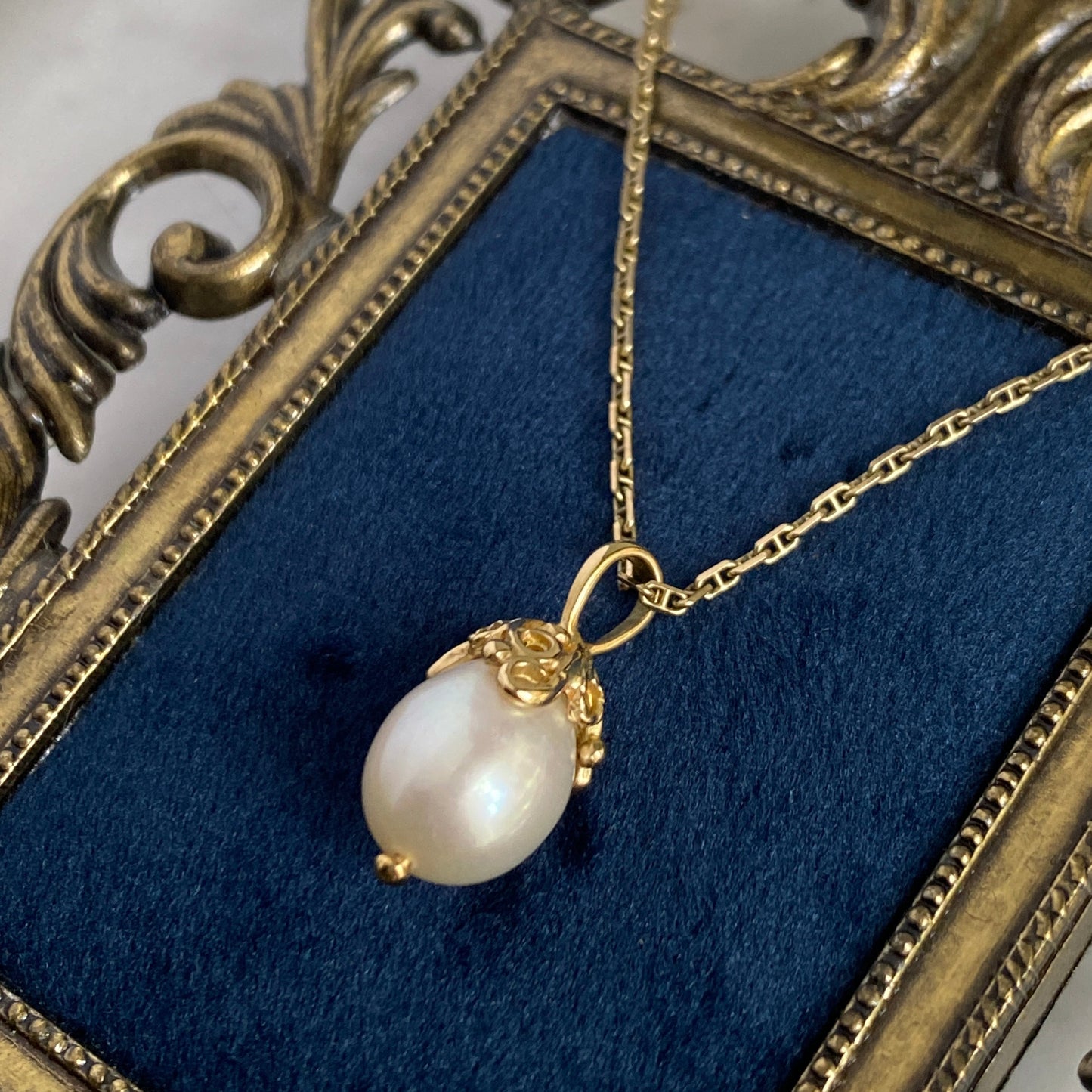 Pearl pendant with a cap of 18k gold filigree swirls on a gold chain in an ornate frame with blue velvet