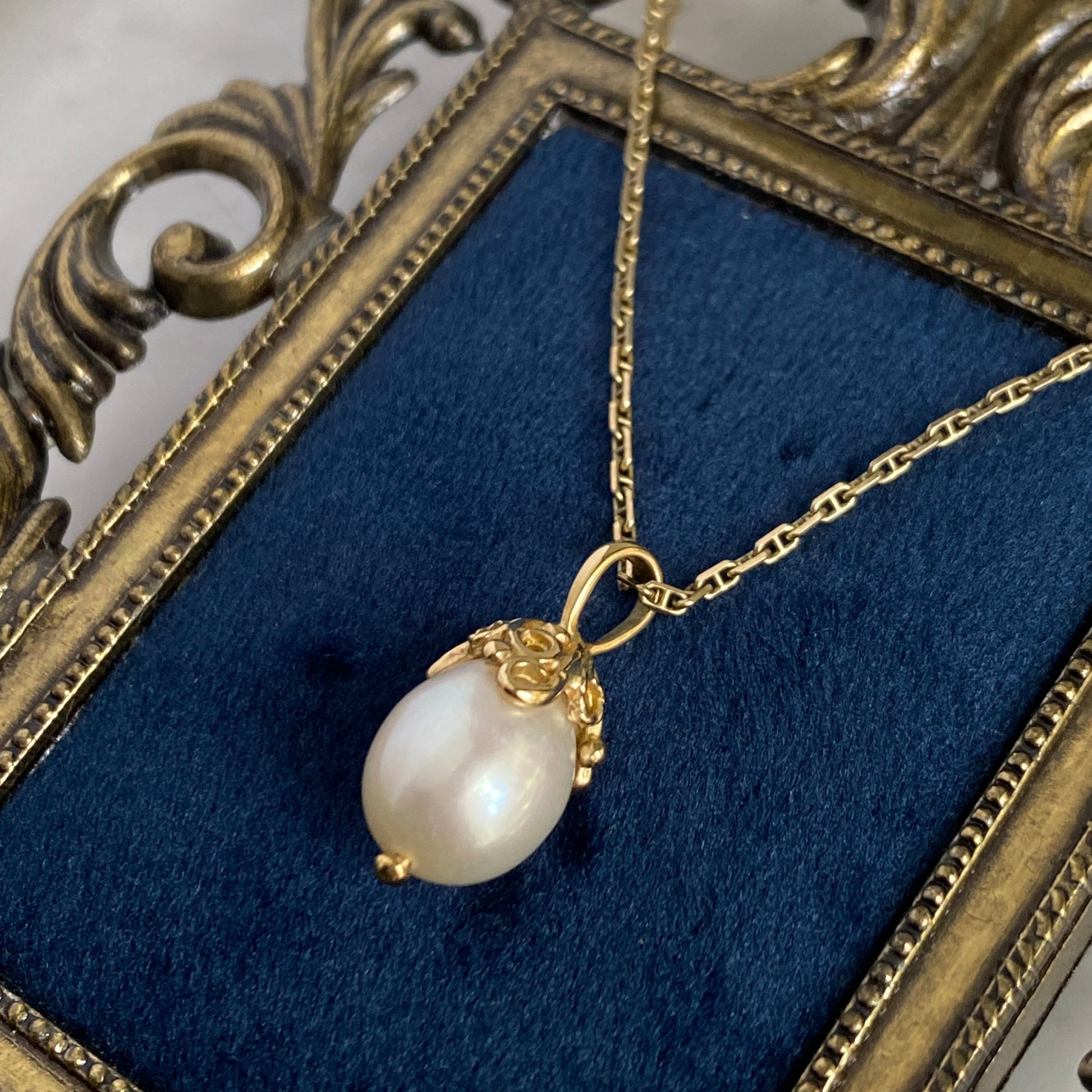 Pearl pendant with a cap of 18k gold filigree swirls on a gold chain in an ornate frame with blue velvet
