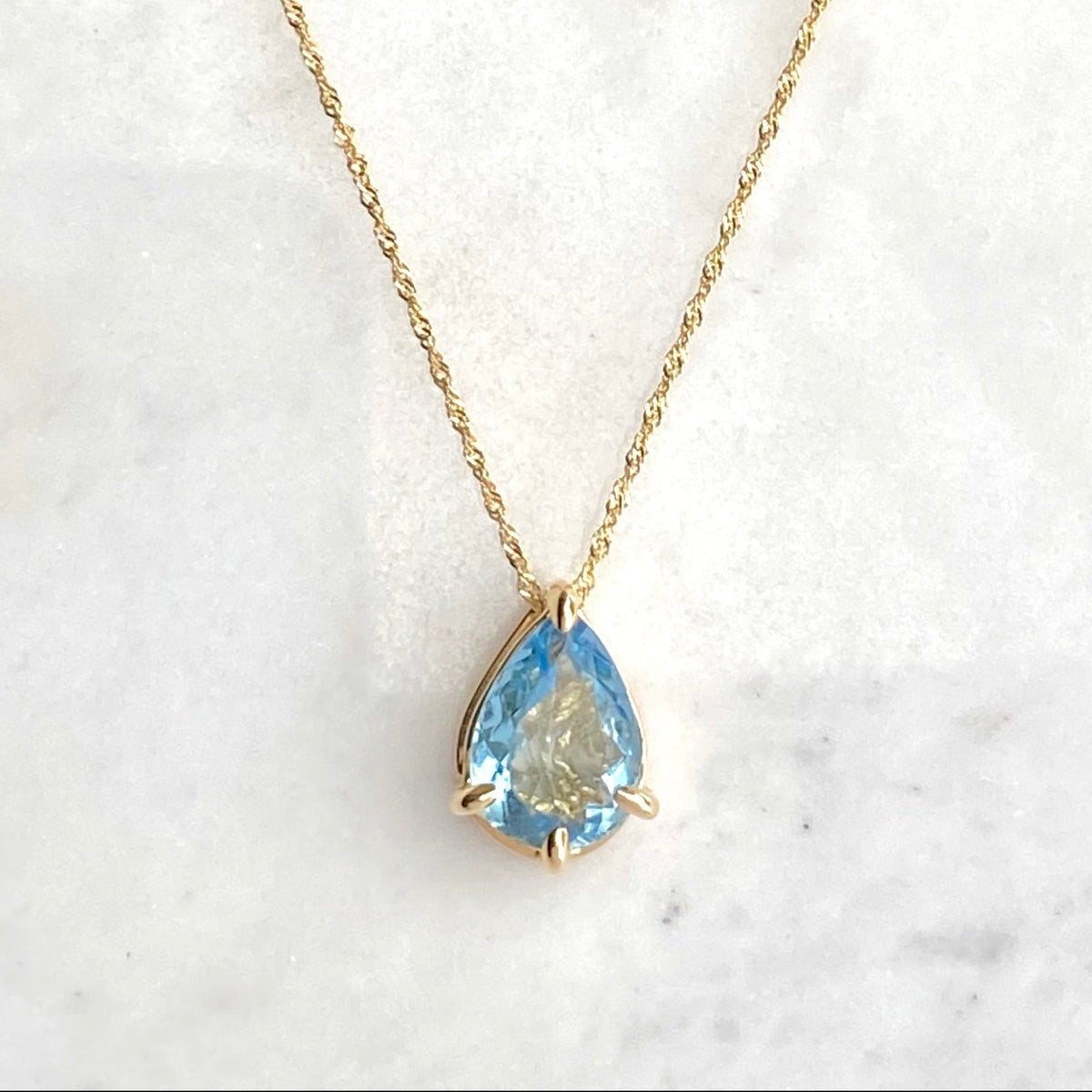 A gold necklace with a pear-shaped light blue aquamarine gemstone pendant, displayed on a white marble surface.