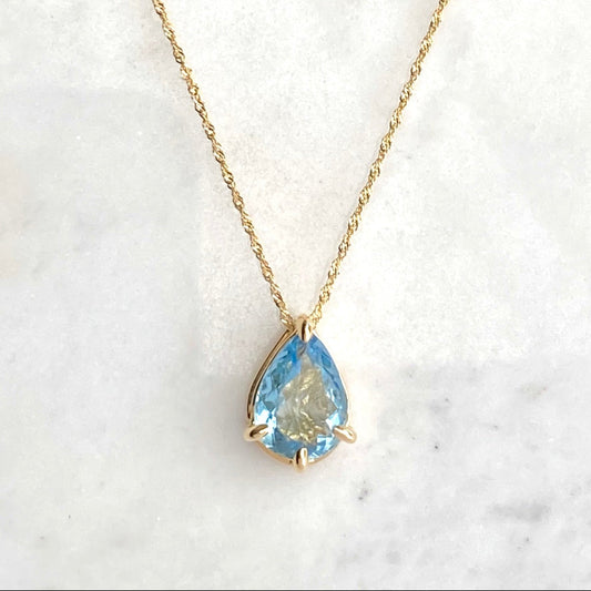 A gold necklace with a pear-shaped light blue aquamarine gemstone pendant, displayed on a white marble surface.