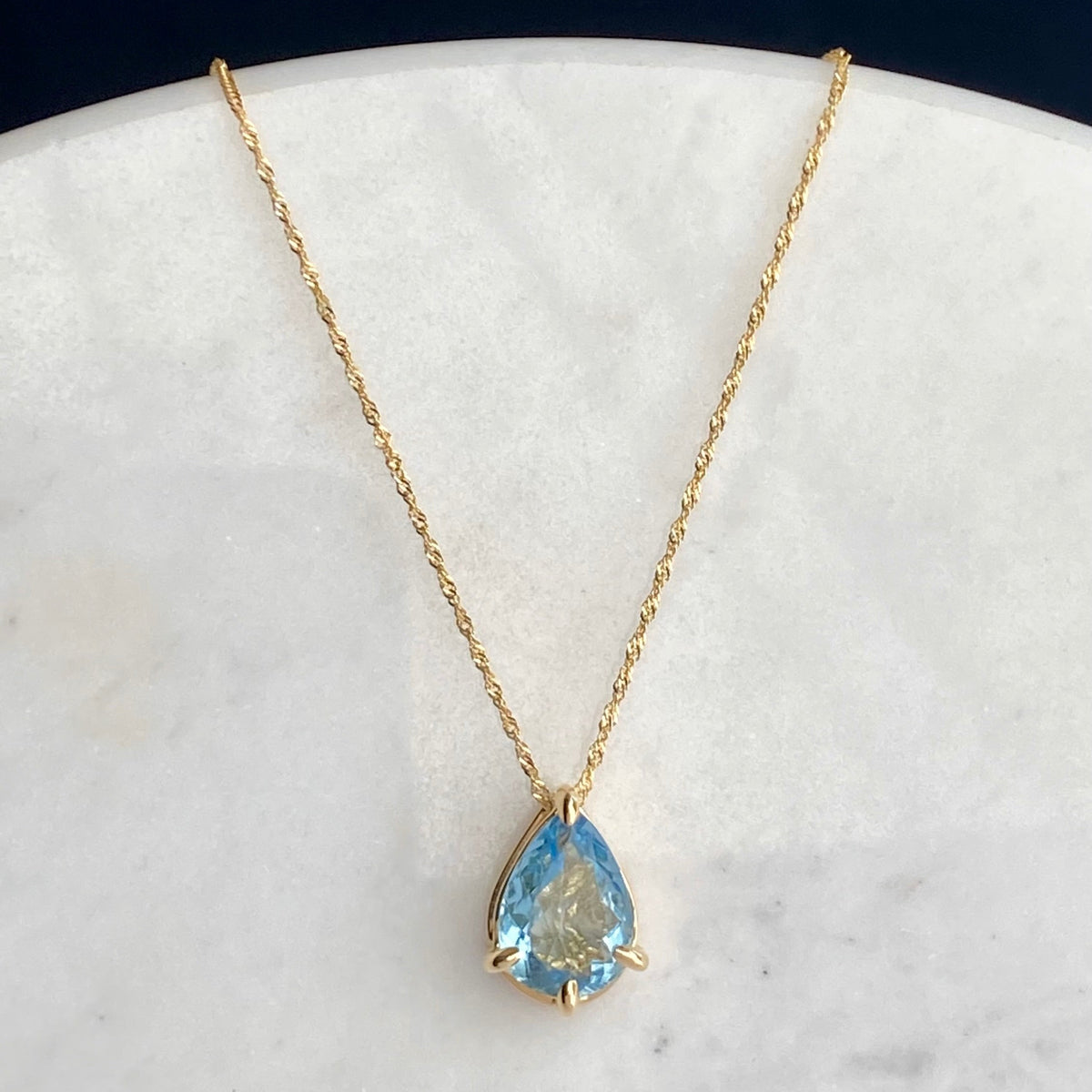 A gold chain necklace with a teardrop-shaped light blue aquamarine gemstone pendant, displayed on a white marble surface against a dark navy background.