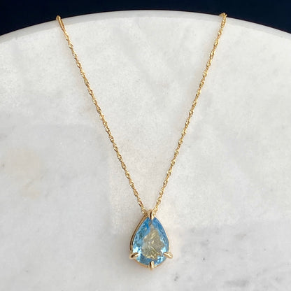 A gold chain necklace with a teardrop-shaped light blue aquamarine gemstone pendant, displayed on a white marble surface against a dark navy background.