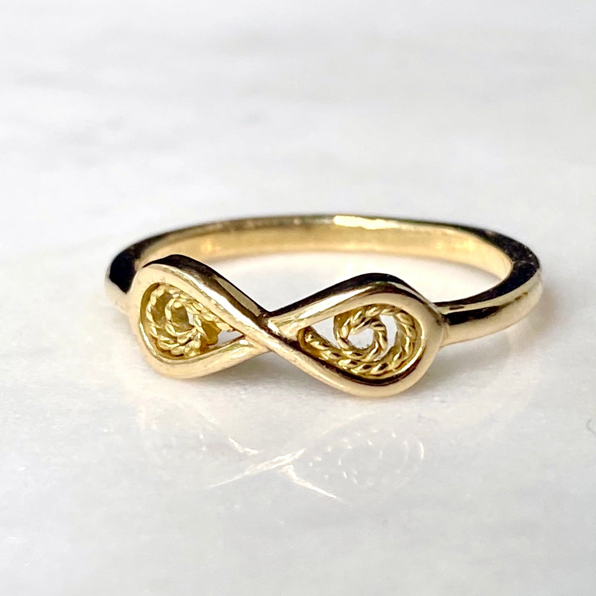 A gold infinity ring with textured rope-like details in the loops, resting on a white marble surface.