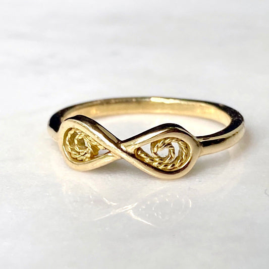 A gold infinity ring with textured rope-like details in the loops, resting on a white marble surface.