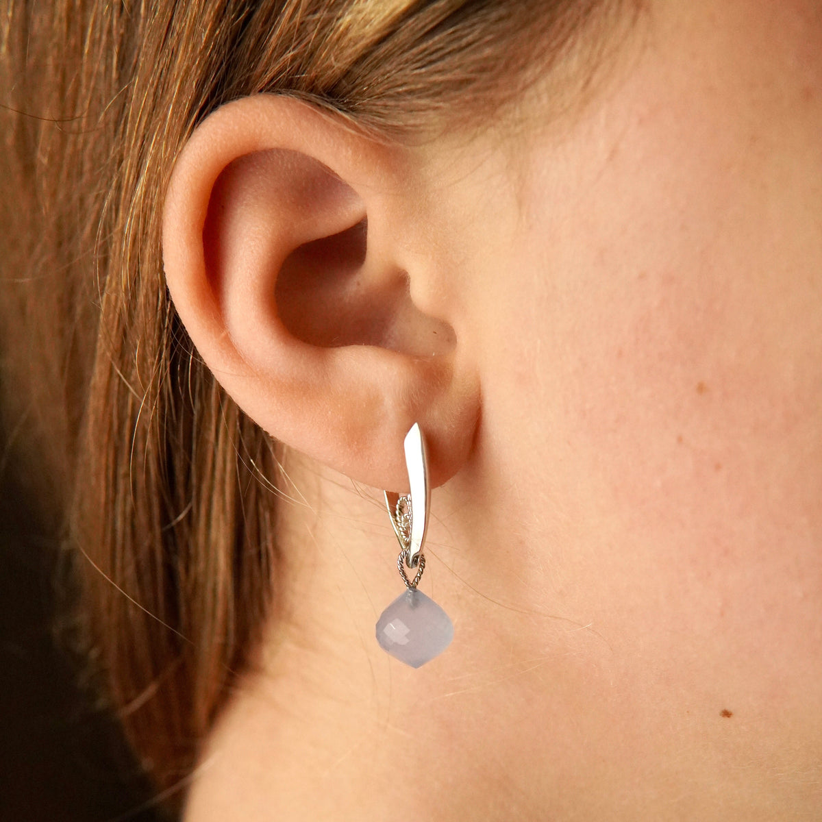 A close-up of a person's ear wearing a silver huggie hoop earring with a small faceted pale blue-gray bead dangle.