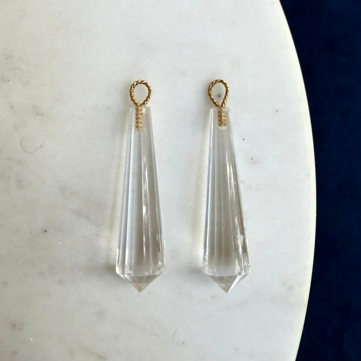 A pair of clear faceted crystal drop earrings with twisted 18k gold loop tops, displayed on a white marble surface.