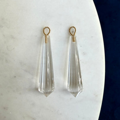 A pair of clear faceted crystal drop earrings with twisted 18k gold loop tops, displayed on a white marble surface.