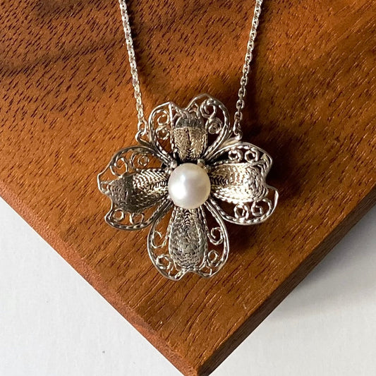 A silver filigree flower pendant necklace with a central white pearl, displayed on a textured brown wooden surface.