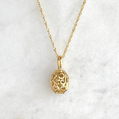 A delicate gold chain necklace with an ornate, filigree-style egg shaped pendant, displayed on a white marble surface against a dark navy blue background.