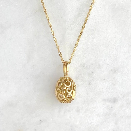 A delicate gold chain necklace with an ornate, filigree-style egg shaped pendant, displayed on a white marble surface against a dark navy blue background.