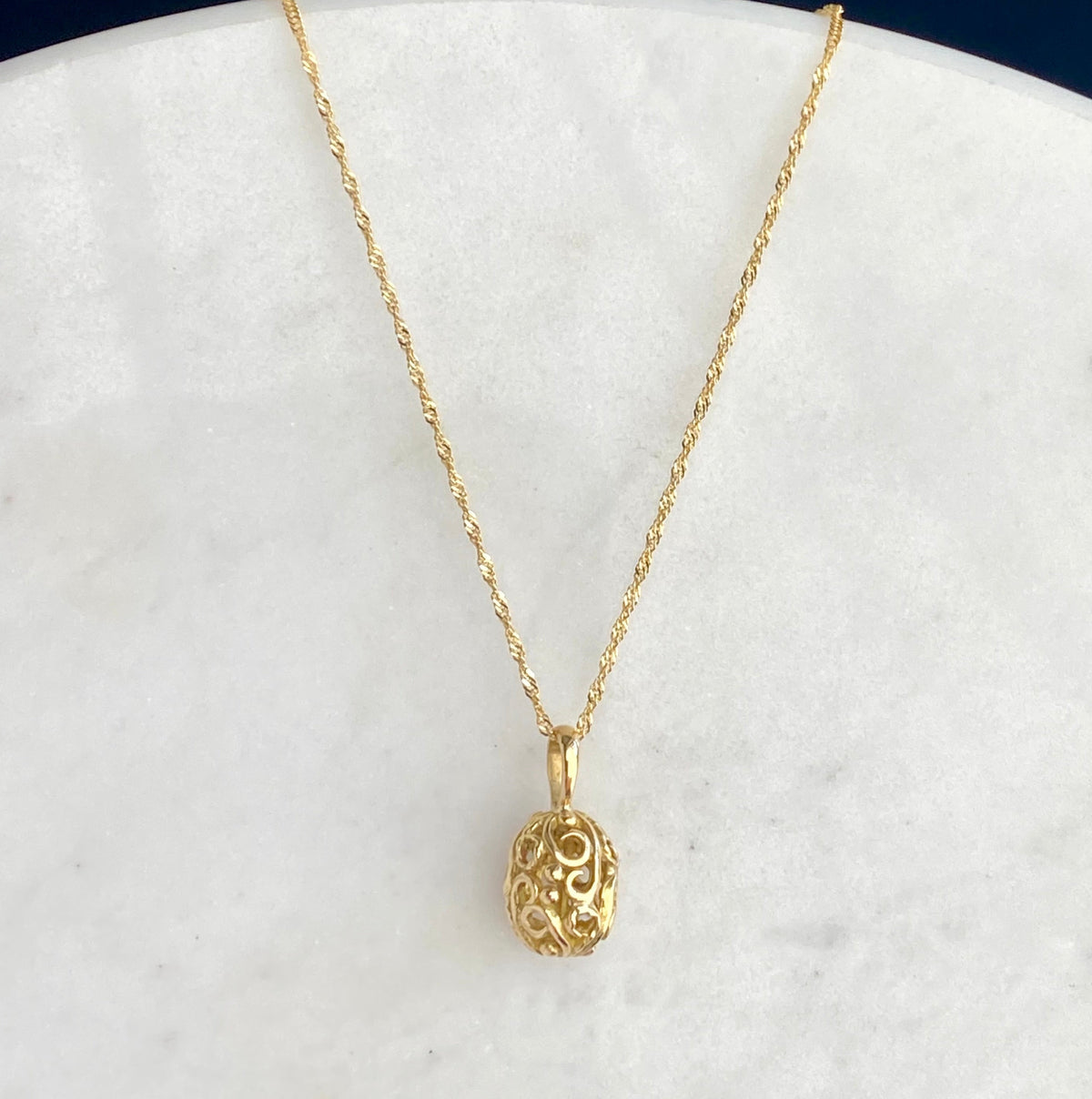 A delicate gold chain necklace with an ornate, filigree egg-shaped pendant, displayed on a white marble surface.