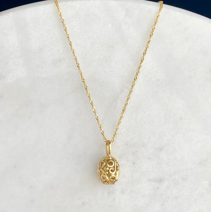 A delicate gold chain necklace with an ornate, filigree egg-shaped pendant, displayed on a white marble surface.
