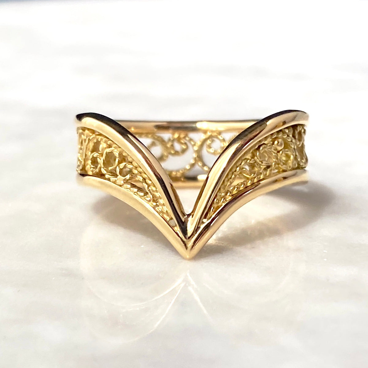 A gold filigree ring with a V-shaped chevron design, resting on a white marble surface.