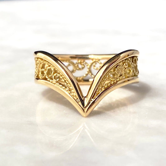A gold filigree ring with a V-shaped chevron design, resting on a white marble surface.
