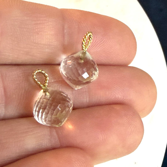 Two small clear faceted onion shaped baubles with gold wire loops, held on a person's fingers against a white and dark background.