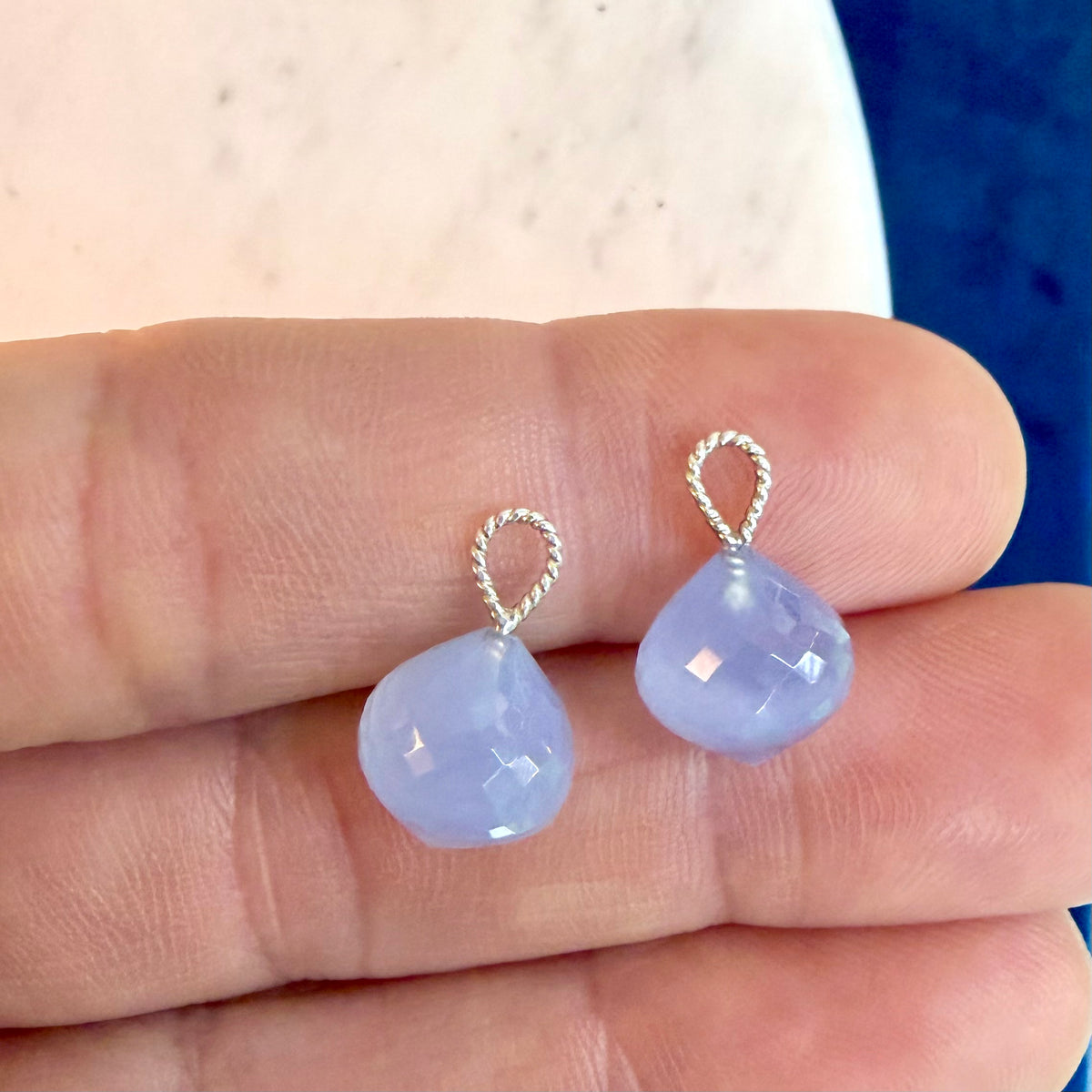 A pair of small teardrop-shaped light blue gemstone drops with silver twisted wire loops, shown resting on a person's fingers.