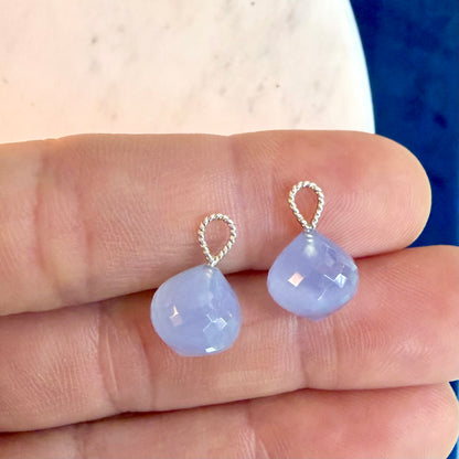 A pair of small teardrop-shaped light blue gemstone drops with silver twisted wire loops, shown resting on a person's fingers.