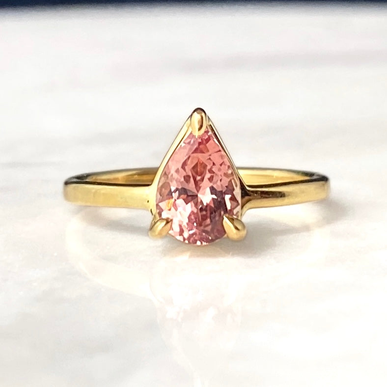 A gold ring featuring a pear-cut pink faceted garnet gemstone in a prong setting, displayed on a light marble surface.