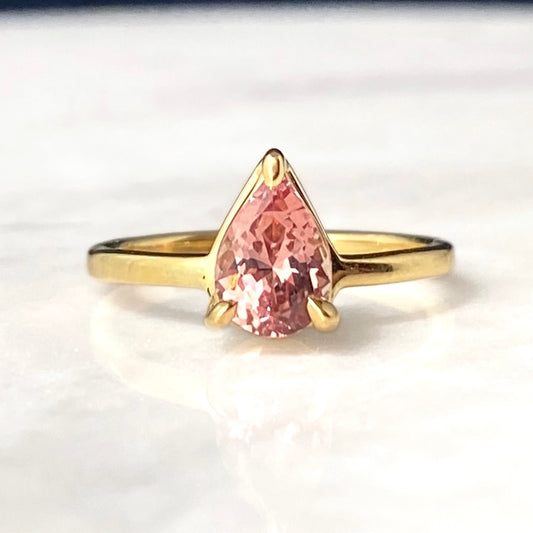 A gold ring featuring a pear-cut pink faceted garnet gemstone in a prong setting, displayed on a light marble surface.