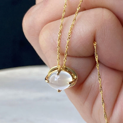 A gold necklace with an oval cats eye moonstone gemstone pendant, held in a person's hand against a light marble surface.