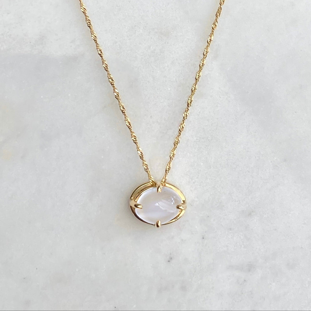 A delicate gold chain necklace with a small oval pendant featuring a milky white cats-eye moonstone stone set in four prongs, displayed on a white marble surface against a dark navy background.