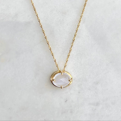 A delicate gold chain necklace with a small oval pendant featuring a milky white cats-eye moonstone stone set in four prongs, displayed on a white marble surface against a dark navy background.