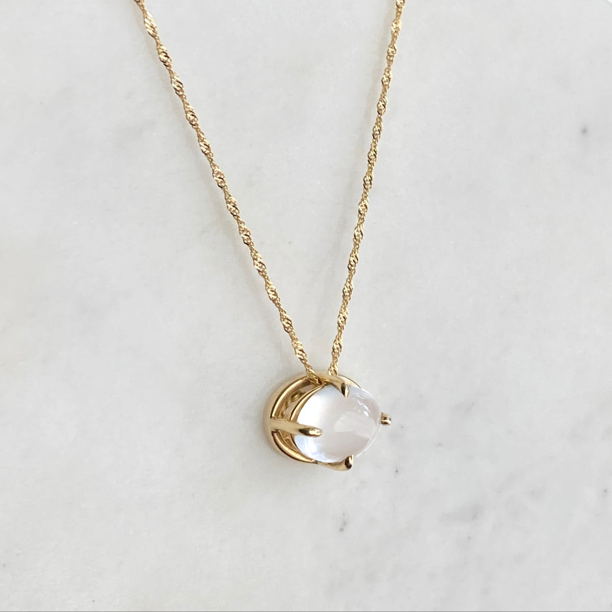 A delicate gold chain necklace with a small gold pendant holding an oval cats-eye moonstone stone, displayed on a white marble surface against a dark blue background.