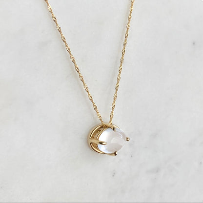 A delicate gold chain necklace with a small gold pendant holding an oval cats-eye moonstone stone, displayed on a white marble surface against a dark blue background.