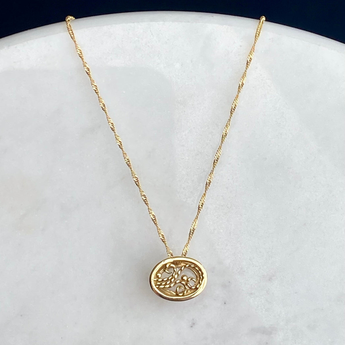 A delicate gold chain necklace with the back of an oval pendant featuring an intricate filigree design, displayed on a white marble surface against a dark navy background.