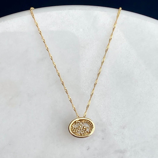 A delicate gold chain necklace with the back of an oval pendant featuring an intricate filigree design, displayed on a white marble surface against a dark navy background.