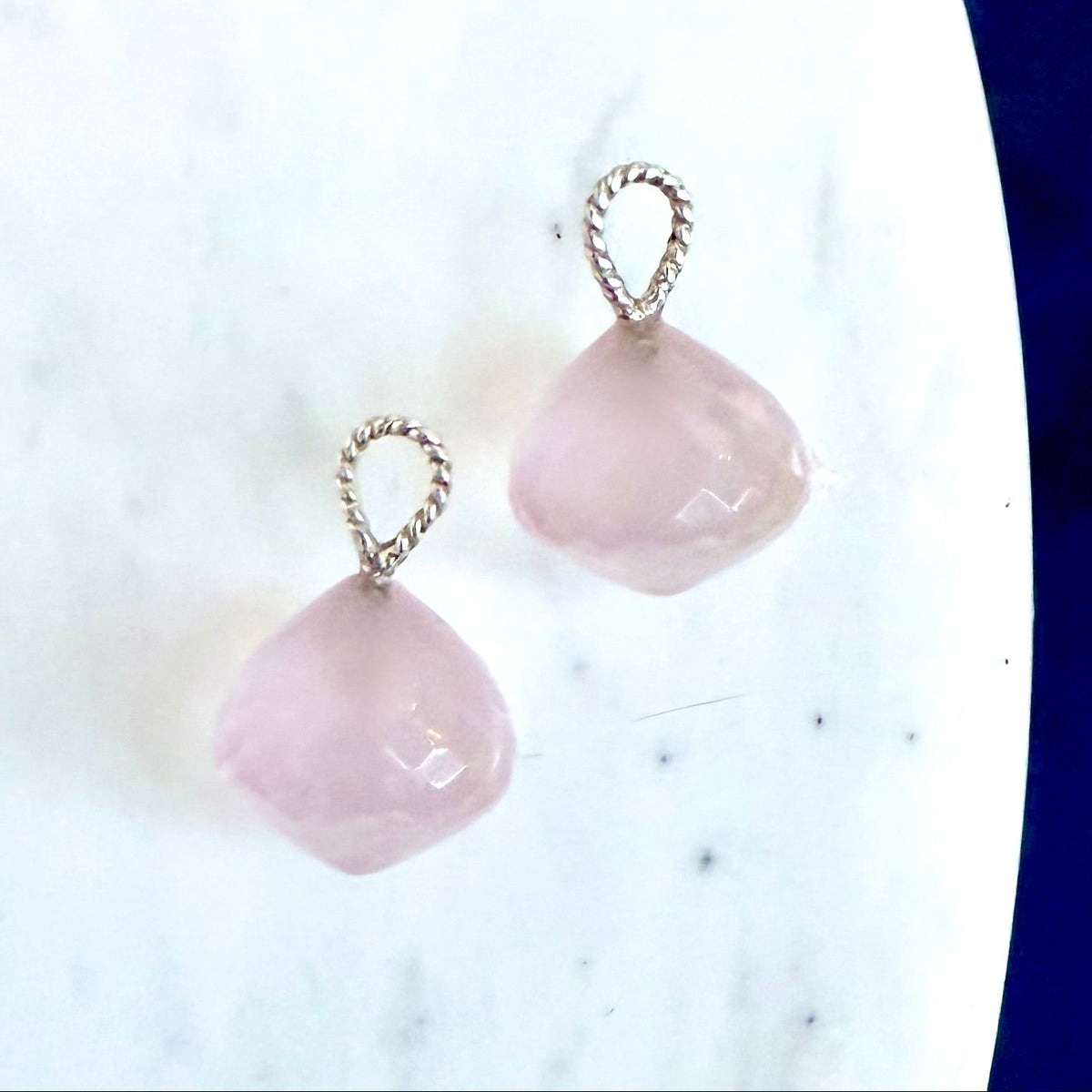 A pair of rose quartz drops with twisted silver wire loops, displayed on a white marble surface with a dark blue background visible on the right.