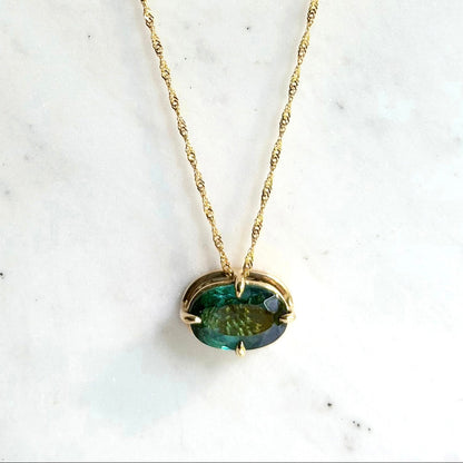 A gold necklace with a twisted chain and an oval blue-green tourmaline gemstone pendant, displayed on a white marble surface.