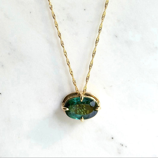 A gold necklace with a twisted chain and an oval blue-green tourmaline gemstone pendant, displayed on a white marble surface.